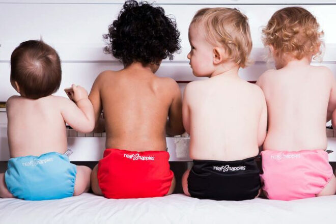 Quatre bébés assis au piano dans des couches en tissu réutilisables Real Nappies montrant les couleurs disponibles